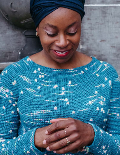 Ginan is a Tunisian jumper with a round neck. In this close-up you can see the increases radiating across the yoke. It uses the assigned pooling method where pops of colour are set against the teal green background.